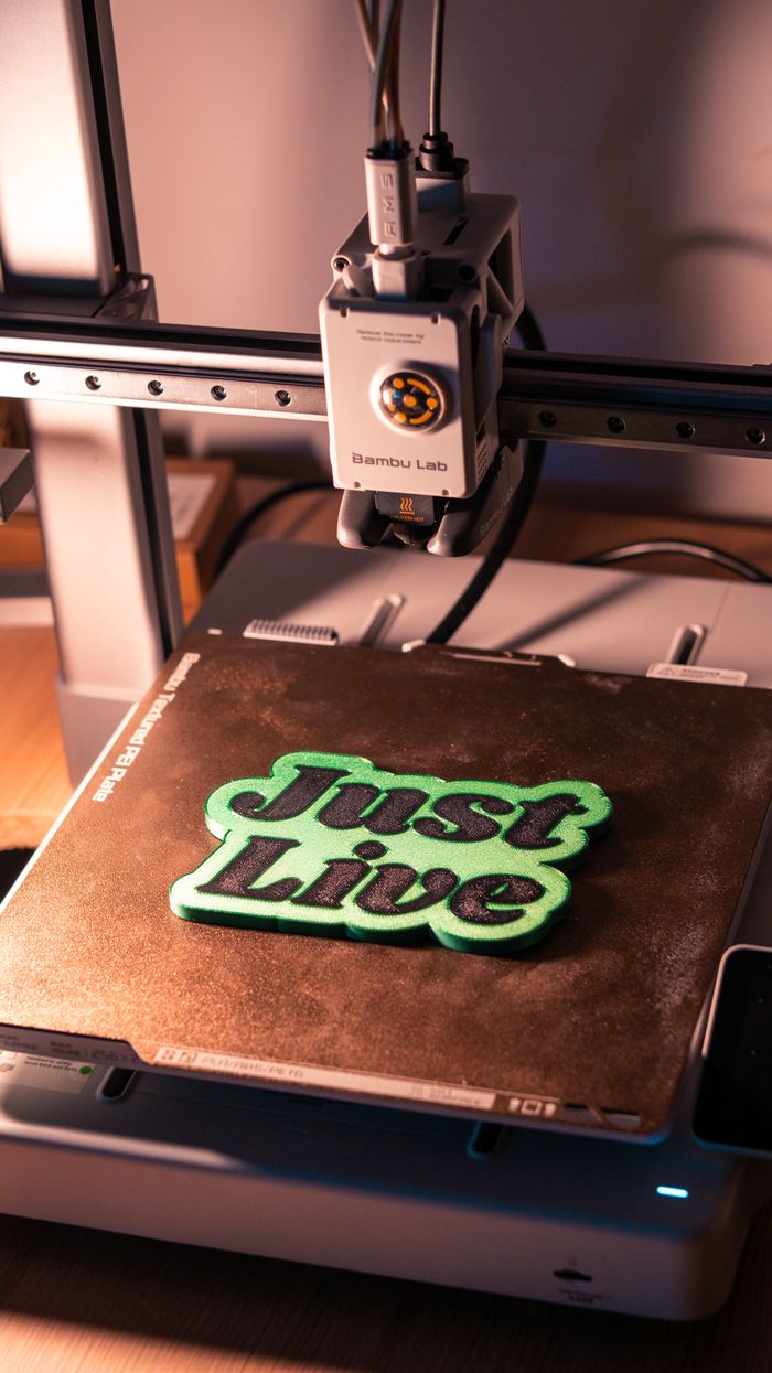 expertise-01 A 3D printer crafting 'Just Live' text in a workshop setting. Ideal for digital technology and innovation themes.